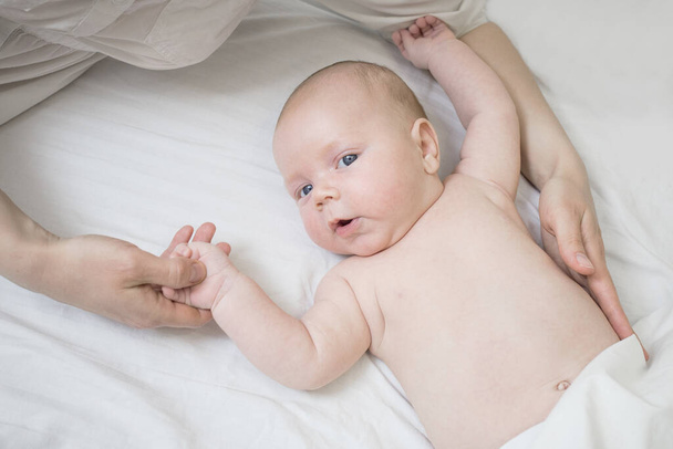 Lieve baby, een blanke pasgeborene, ligt in bed en houdt zijn moeders hand vast. Naakt, bedekt met een witte deken. Het concept van babyverzorging, veiligheid, ouderschap, geluk van het moederschap, liefde.  - Foto, afbeelding