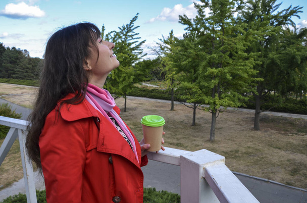 Buiten portret van vrolijke mooie brunette vrouw in rode jas met wegwerp koffiekopje drinken van koffie, en glimlachen tegen stedelijke natuur achtergrond. Koffie te gaan  - Foto, afbeelding