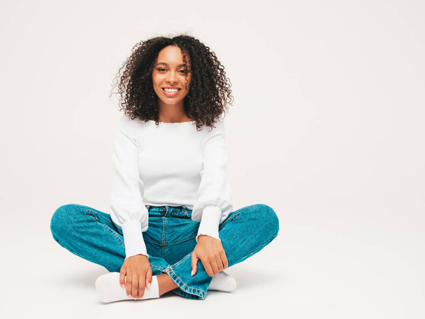 Beautiful black woman with afro curls hairstyle.Smiling model in sweater and trendy jeans clothes. Sexy carefree female sitting on white background in studio. Tanned and cheerful  - Foto, imagen