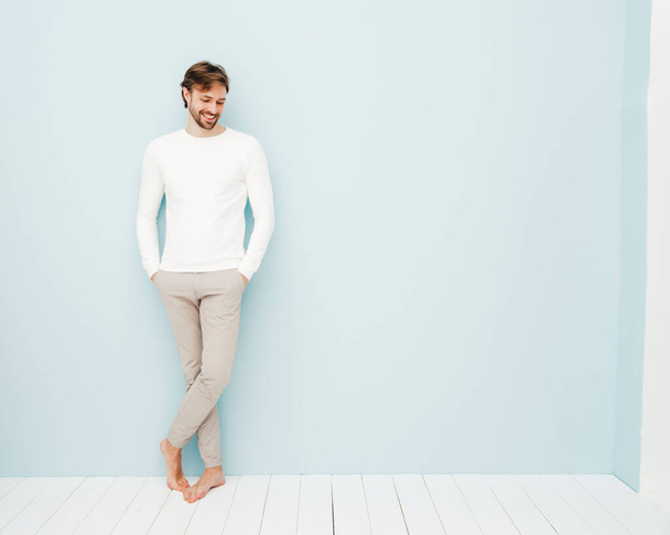 Portrait of handsome smiling hipster lumbersexual businessman model wearing casual white sweater and trousers. Fashion stylish man posing against light blue wall in studio - Foto, afbeelding