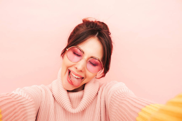 Young beautiful smiling female in trendy summer colorful sweater. Sexy carefree woman posing near pink wall in studio.Positive model having fun in sunglasses. Taking Pov selfie photos.Shows tongue - Foto, imagen
