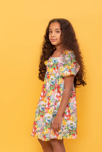 Половина портрета темнокожей молодой девушки с длинными вьющимися волосами в красочном платье на жёлтом фоне - Фото, изображение