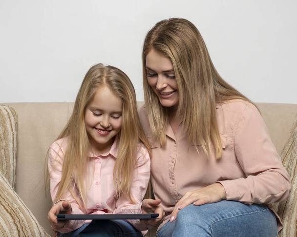 Фотография мамы и дочери блондинок с длинными волосами в розовых рубашках и джинсах, сидящих дома на диване с ноутбуком.   - Фото, изображение