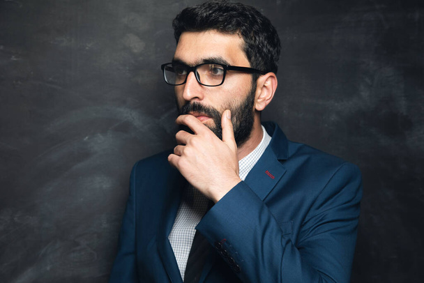 mladý podnikatel stojí a přemýšlí s rukou na bradě na tmavém pozadí - Fotografie, Obrázek