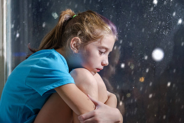 A brown eyed girl in blue t-shirt sitting on the window sill behind a foggy window with snowy night background - Photo, Image