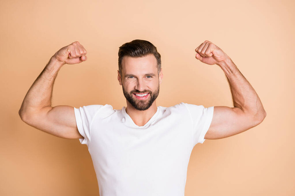 Portrait of attractive cheerful guy showing muscles champion competitor isolated over beige pastel color background - Foto, imagen