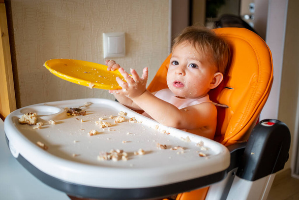 adorable bebé juega con el plato en la mesa. niño pequeño se entrega en una silla de bebé después de comer - Foto, imagen