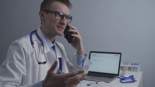 Médico europeo en bata médica blanca y gafas consulta al paciente en el teléfono celular en la oficina de la clínica, sentado en la mesa con el ordenador portátil. Médico general alemán en el lugar de trabajo en el hospital hablando por teléfono - Metraje, vídeo