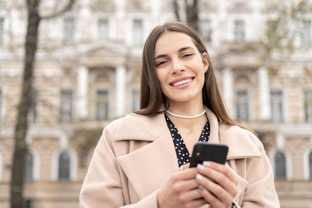 Retrato de una alegre mujer de negocios sosteniendo un teléfono móvil en un parque de la ciudad con una sonrisa perfecta - Foto, Imagen