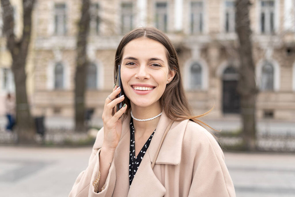 Портрет веселой женщины, разговаривающей по мобильному телефону с широкой улыбкой, стоящей в городском парке  - Фото, изображение