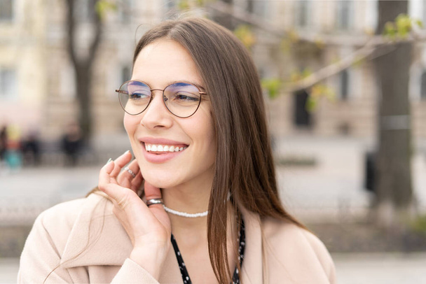 Porträt einer fröhlichen Frau mit Brille und perfektem Lächeln, die im Stadtpark auf die Seite schaut  - Foto, Bild