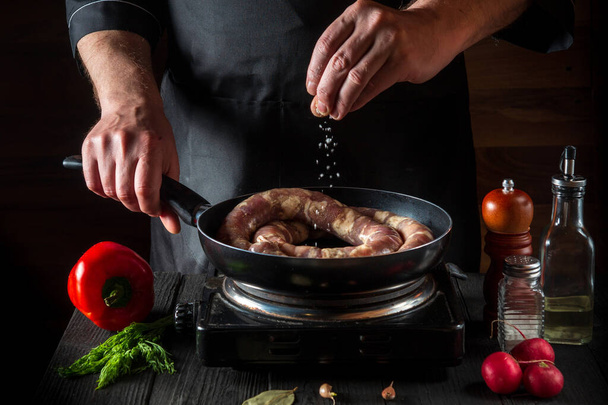 The chef adds salt to pan with raw meat sausage. Preparation for cooking sausages in the kitchen of a restaurant or cafe on a table with vegetables and spices - Foto, afbeelding