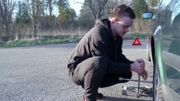 Een jongeman schroeft een wiel los op een auto, hij draagt een zwart trainingspak en zwarte sneakers, een concept van een kapotte auto op de weg. - Video