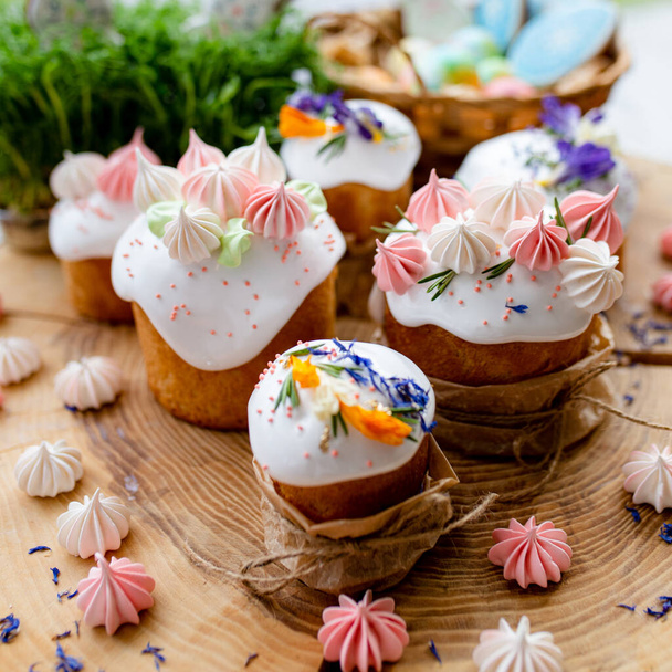 Easter decor 2021. Cakes with white icing, meringue and flowers, gingerbread cakes in icing and colorful painted eggs in a wicker basket on a natural wooden background. Easter card. - Photo, Image