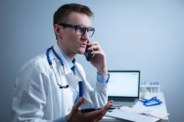 Médico europeo en bata médica blanca y gafas consulta al paciente en el teléfono celular en la oficina de la clínica, sentado en la mesa con el ordenador portátil. Médico general alemán en el lugar de trabajo en el hospital hablando por teléfono. - Foto, imagen