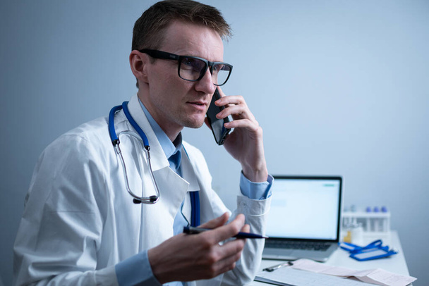 Europäischer Arzt in weißem Arztkittel und Brille berät Patienten per Handy im Klinikbüro, am Tisch mit Laptop sitzend. Deutscher Allgemeinmediziner am Arbeitsplatz im Krankenhaus telefoniert. - Foto, Bild