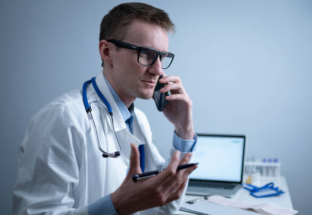 Médico europeo en bata médica blanca y gafas consulta al paciente en el teléfono celular en la oficina de la clínica, sentado en la mesa con el ordenador portátil. Médico general alemán en el lugar de trabajo en el hospital hablando por teléfono. - Foto, imagen
