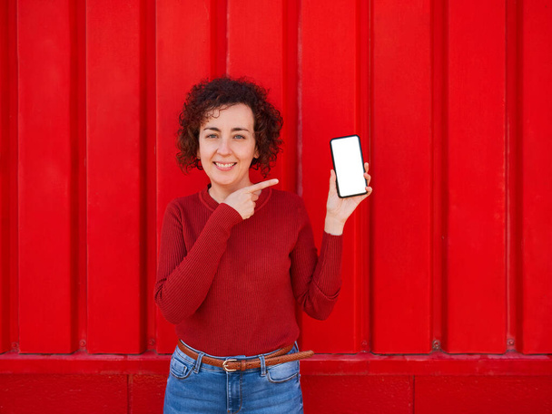 Egy fiatal fehér nő göndör hajjal megmutatja a telefont a kamerának, miközben mosolyog, piros pulóvert és farmert visel, és a háttérben van egy vörös fal, nappal van.. - Fotó, kép