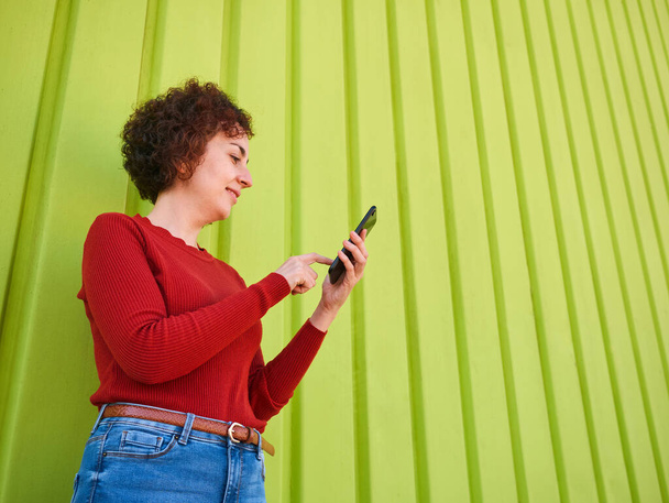 Una giovane donna caucasica con i capelli ricci che indossa un maglione rosso e jeans usa il suo cellulare per navigare in internet, ha un muro verde sullo sfondo ed è giorno - Foto, immagini