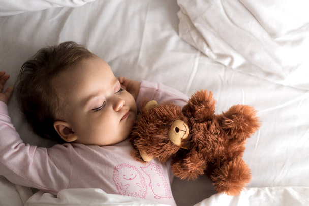 Authentisches Porträt niedlichen kaukasischen kleinen Säugling pummelig Baby Mädchen oder Junge im rosa Schlaf mit Teddybär auf weißem Bett. Mittags ruhen die Kinder. Pflege, Schlafkind, Kindheit, Elternschaft, Lebenskonzept - Foto, Bild