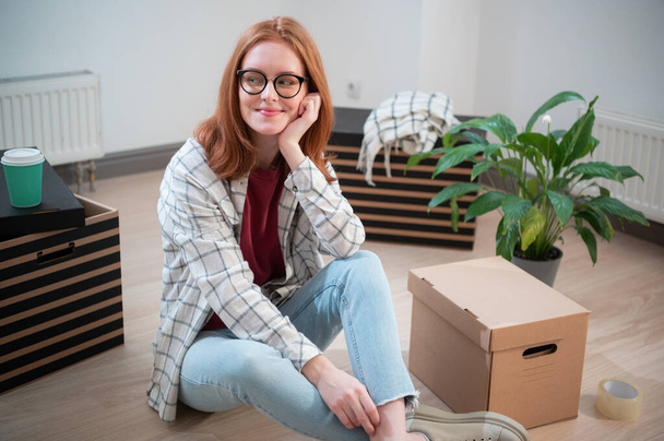 A pretty young woman with glasses is tired when moving to a new apartment, she sits on the floor next to boxes and a potted plant and dreams - 写真・画像