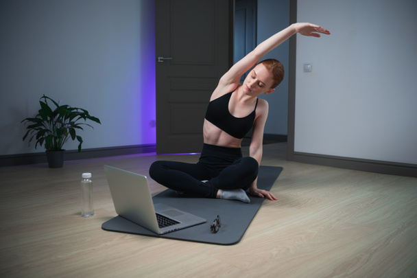 Stretching exercises are an important part of online training in the course on body balance, the girl is engaged on the mat at home in front of the laptop - Photo, Image