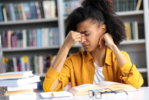 Cansada estudiante afroamericana agotada sentada en una mesa con libros en la biblioteca, cerró los ojos, masajeando el puente de su nariz, dolor de cabeza y estrés por el exceso de trabajo de aprender - Foto, Imagen