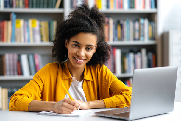 Afrikkalaisen teinitytön muotokuva. Miellyttävä sekarotuinen naisopiskelija istuu kirjastossa kirjoituspöydän ääressä kannettavan tietokoneen kanssa, tekee muistiinpanoja luennon aikana, katsoo kameraa, hymyilee - Valokuva, kuva