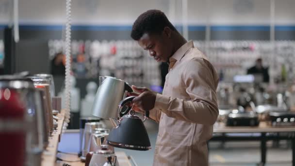jonge zwarte man vergelijkt twee elektrische waterkoker in de winkel, het houden van zowel in de handen en wegen, het bekijken van verschillende modellen - Video