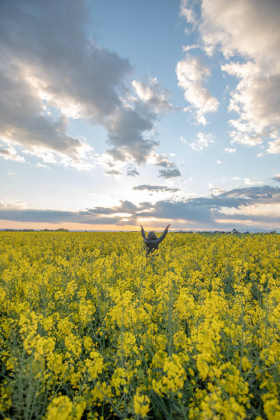 mujer con los brazos levantados con un anorak negro con la capucha en un campo de flores de colza amarillas al atardecer con nubes en el cielo, vertical - Foto, Imagen