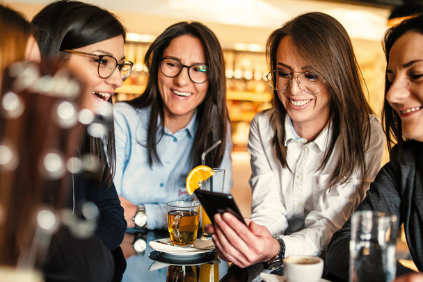 Seitenansicht einer erwachsenen kaukasischen Frau mit Brille, die ihr Handy in der Hand hält, während sie mit ihren Millennial-Freundinnen im Café oder Restaurant sitzt und ein Videotelefonat auf dem Smartphone führt oder Selfie-Fotos macht - Foto, Bild