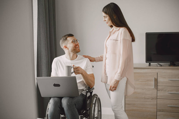 Young man in wheelchair using laptop and girlfriend hugging him at home - Φωτογραφία, εικόνα