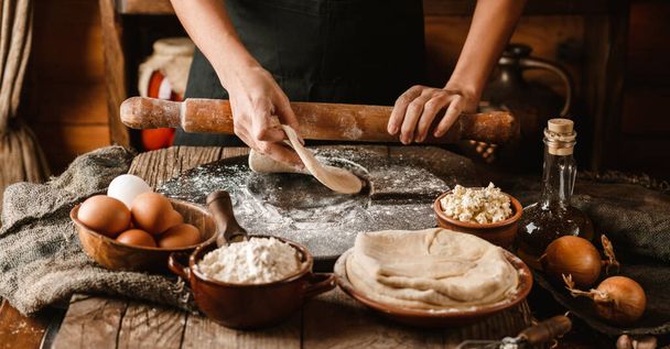 Woman hands rolls the dough with a rolling pin on rustic wooden background. Cooking bread with cheese, eggs and herb. Homemade healthy food concept - Photo, Image