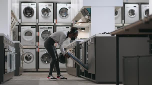 jovem mulher negra com máscara facial está vendo máquina de lavar louça na loja, shopping com departamento de eletrodomésticos, escolhendo equipamentos para cozinha - Filmagem, Vídeo