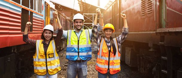 Inżynierowie logistyki kolejowej lub pracownicy przemysłu w kamizelkach bezpieczeństwa i kaskach podnosili pięść, aby pokazać radość i pewność siebie na zewnątrz między lokomotywami.. - Zdjęcie, obraz