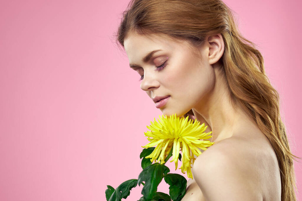Mooie vrouw met naakte schouders en een gele bloem in haar handen op een roze achtergrond - Foto, afbeelding