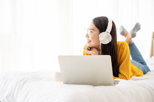 Hermosa mujer joven asiática que usa auriculares y computadora portátil. Ella ocio en la cama en el dormitorio - Foto, imagen