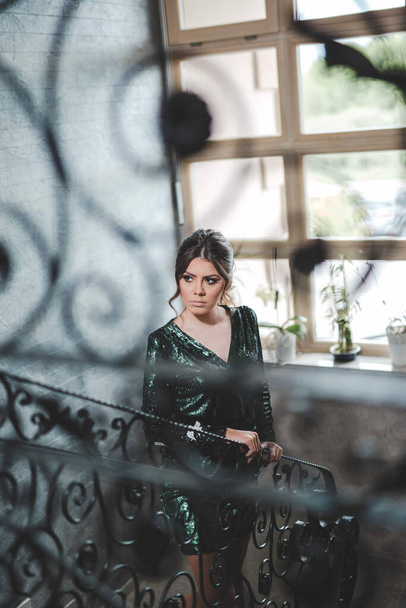 A sexy Bosnian brunette girl in stylish green sequins dress from behind a black decorative stair fence - Foto, afbeelding