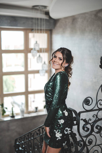 A vertical shot of a beautiful Caucasian woman in a shiny green dress, posing indoors in a decorative stairwell - Fotografie, Obrázek