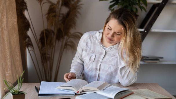Portrait of a Caucasian teacher sitting at a wooden table with books and textbooks at home by the window. The woman is preparing for the lesson. Homeschooling. - Photo, Image