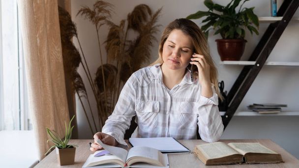Πορτρέτο μιας νεαρής καυκάσιας γυναίκας συμβούλου γιατρού που κάθεται σε ένα ξύλινο τραπέζι με βιβλία και εγχειρίδια στο γραφείο δίπλα στο παράθυρο. Η γυναίκα μιλάει στο τηλέφωνο και διαβάζει λογοτεχνία.. - Φωτογραφία, εικόνα