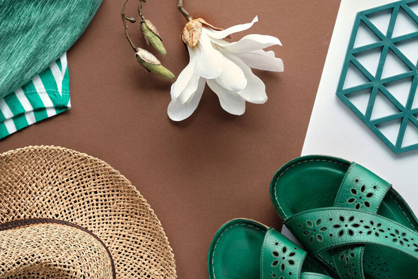 Natural trendy spring female fashion outfit. Flat lay, top view on split two tone paper geometric background. White magnolia flower and hexagones. Straw hat, tshirts, green leather sandals. - Фото, изображение
