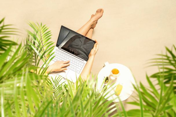 Woman freelancer with laptop sitting under palm tree branches. Sunscreen, sunglasses, orange juice on the table of sandy beach . Summer vacation. Top view - Foto, immagini