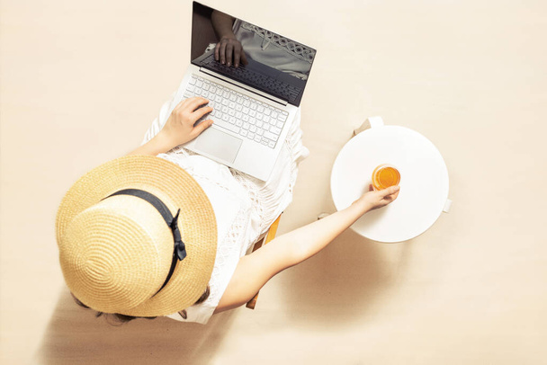 Woman freelancer with laptop holding glass of water with piece orange sitting under palm tree branches on the sand of beach. Outdoors cafe. Summer vacation. Top view - Fotografie, Obrázek