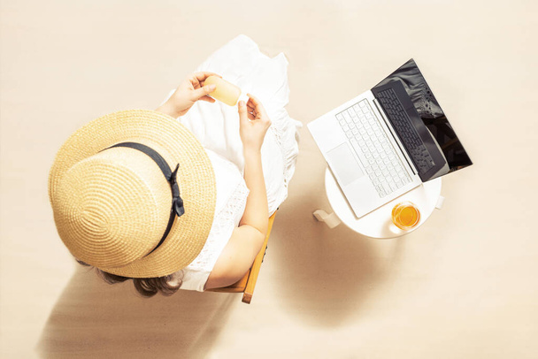 Woman blogger with laptop applying sunscreen on skin sitting near table on the sand of beach . Summer vacation. Top vie - Fotoğraf, Görsel