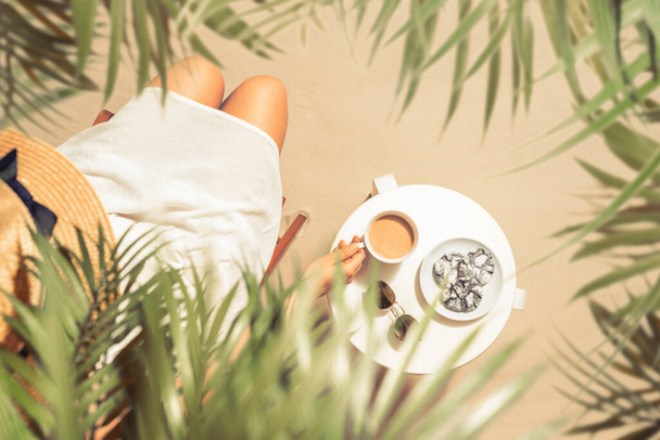 Woman on the beach drinking hot coffee with chocolate cookies sitting under palm trees at table and enjoying during exotic summer vacation. Outdoors cafe. Top view - Fotoğraf, Görsel