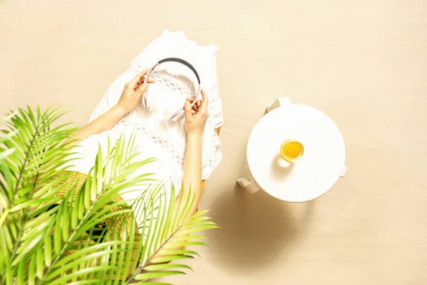 Alone woman sitting under palm tree branches. Glass of orange juice and headphones on the table. Female relaxation on the sandy beach at summer vacation. Top view - 写真・画像