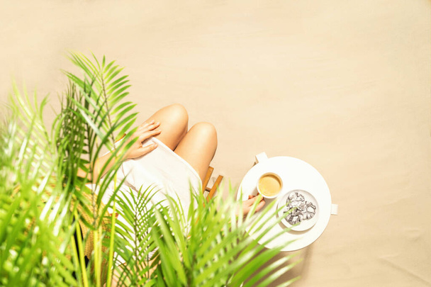 Woman drinking hot coffee with chocolate cookies sitting under palm trees at table. Female on the beach enjoying exotic summer vacation. Outdoors cafe. Top view - Photo, image