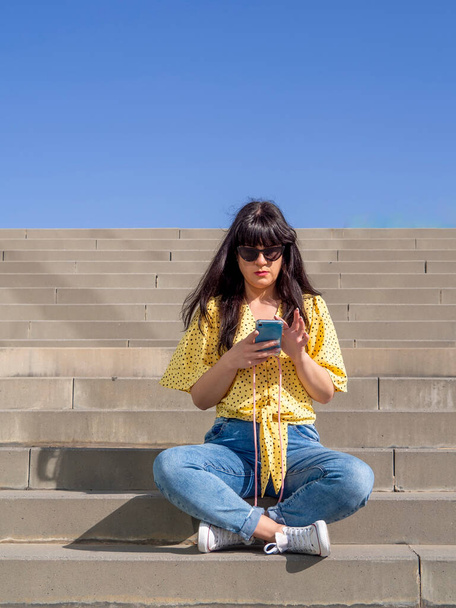 Mujer joven morena sentada en las escaleras afuera y usando un teléfono celular en un día de verano. Chica con gafas de sol de mensajería con teléfono inteligente serio. Adicción a las redes sociales - Foto, Imagen