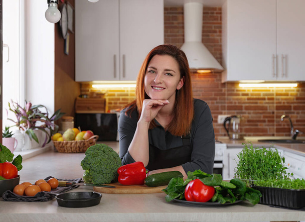 Концепция здорового питания портрет красивой молодой рыжеволосой женщины, готовящей салат с брокколи, цуккини, красным перцем, травами на столе - Фото, изображение